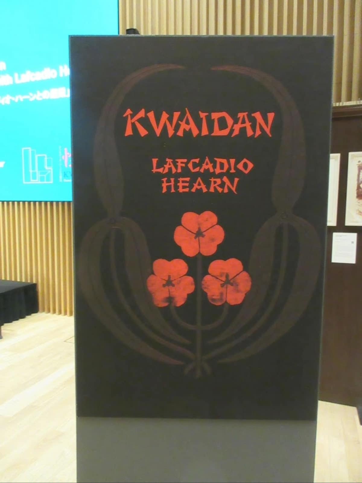 Official title board for the "Kwaidan – Encounters with Lafcadio Hearn" exhibition featuring a red floral crest on a black background.
