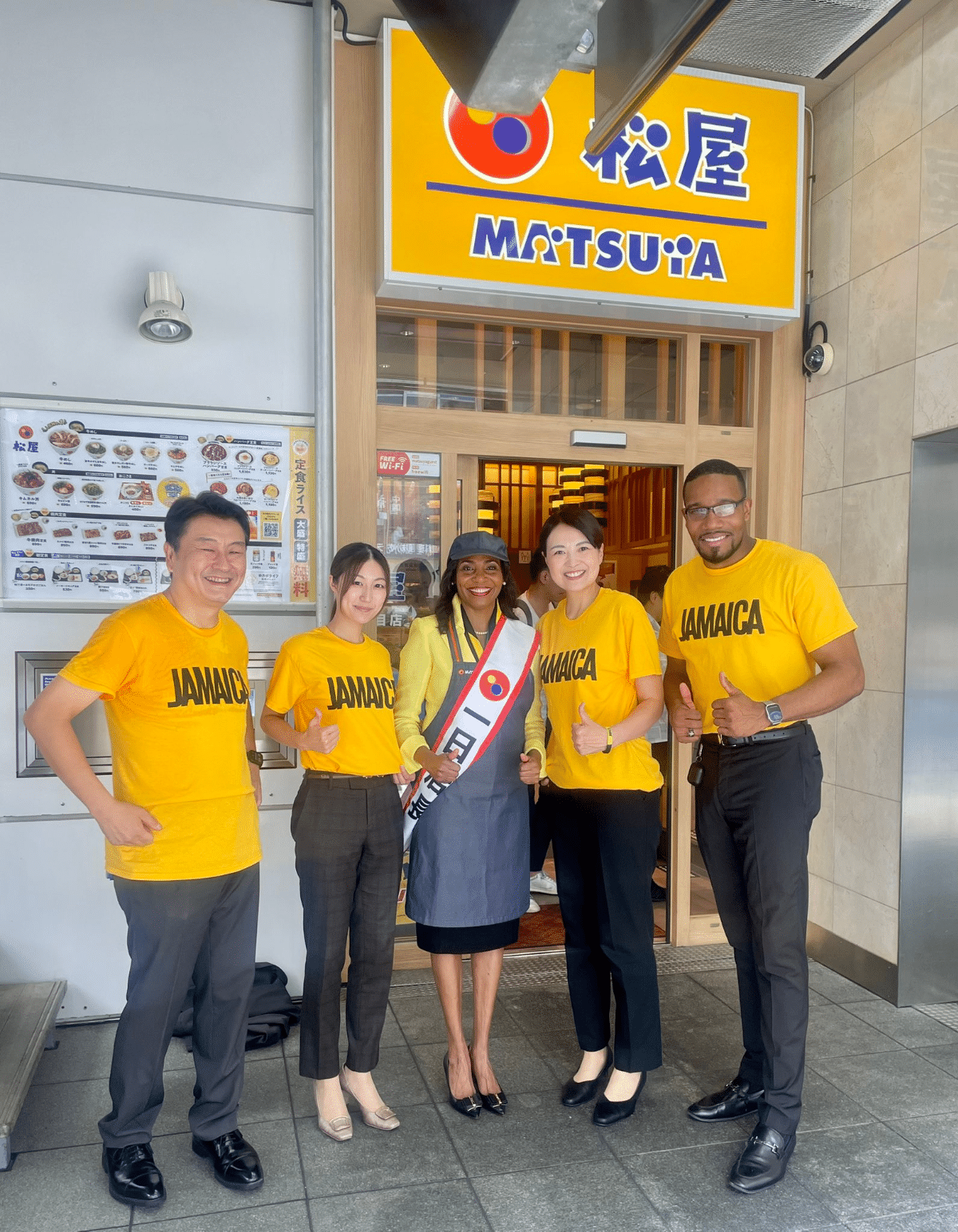 松屋六本木店で「一日店長」を務めるショーン・ケイ・リチャーズ大使。ジャマイカのTシャツを着たスタッフと共に、ジャマイカ料理のジャークチキンをPR。