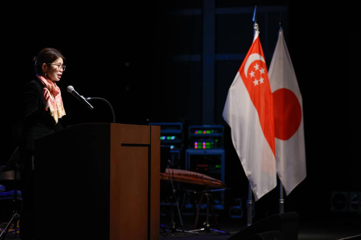 Esther Ong speaking at the Singapore Embassy event in Tokyo, with the flags of Singapore and Japan in the background.