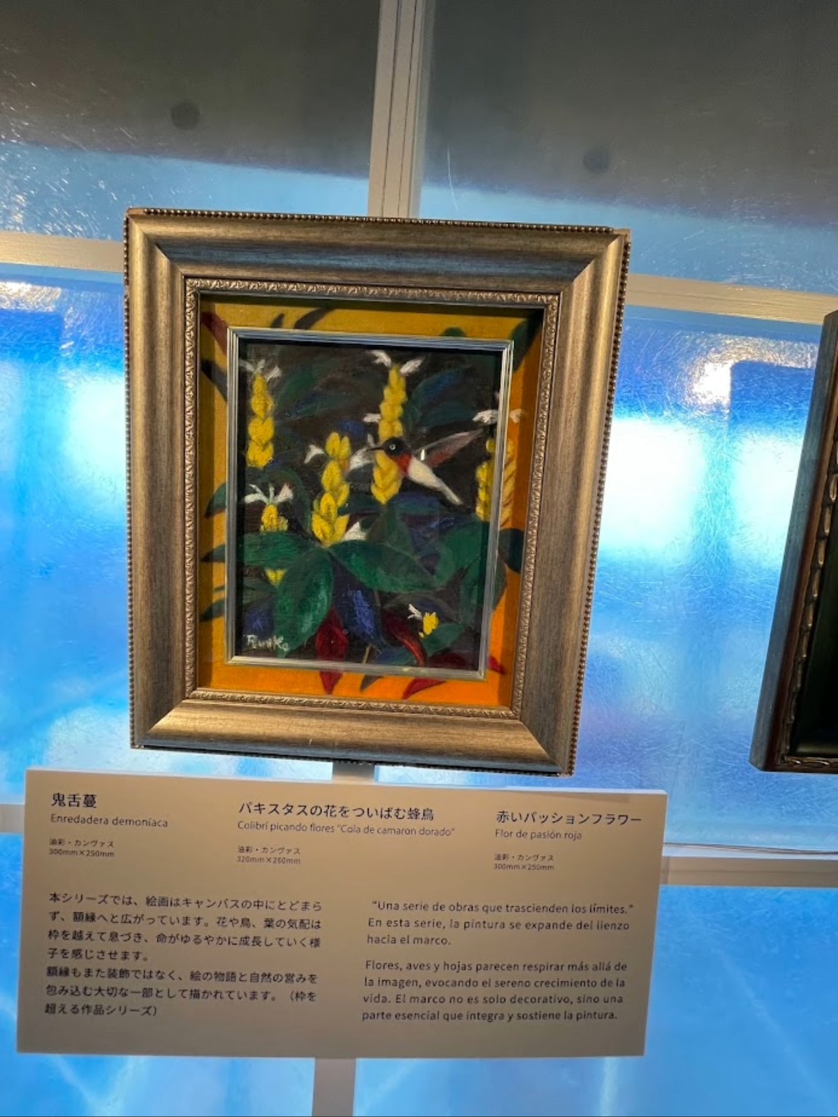 額縁を超えて描かれたハチドリの作品と、会場の解説パネル。