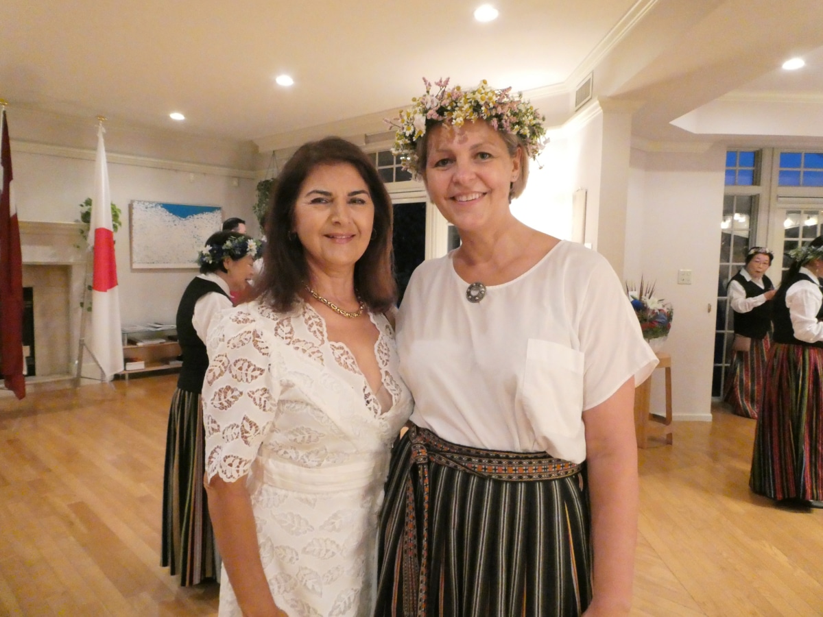 Madam Ilona Zilgalve, wearing a floral crown, poses with a guest at the farewell reception, with Choir Gaisma members in folk costume nearby.