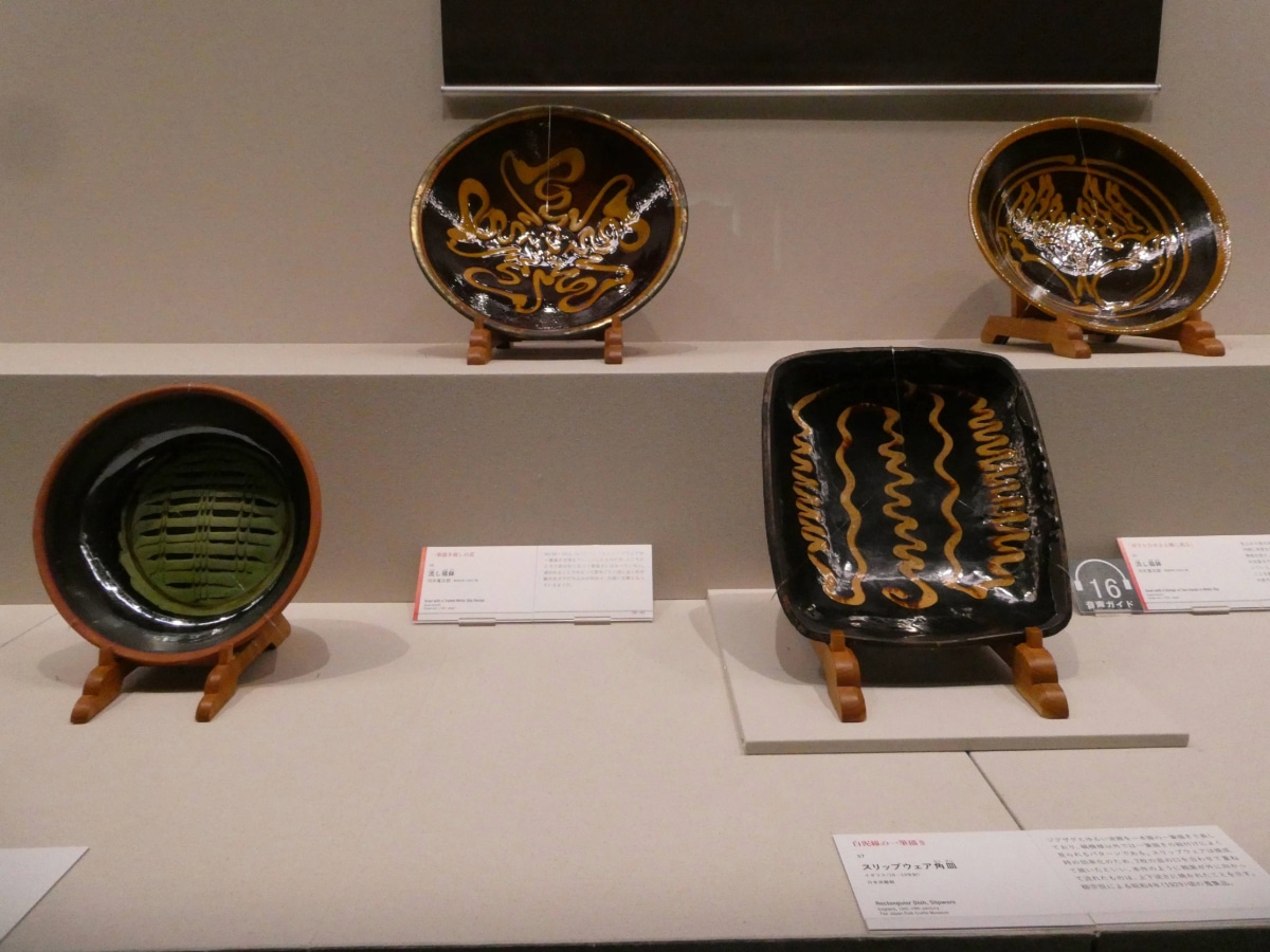 English slipware dishes with wavy yellow slip patterns on dark glaze, shown in the Mingei section