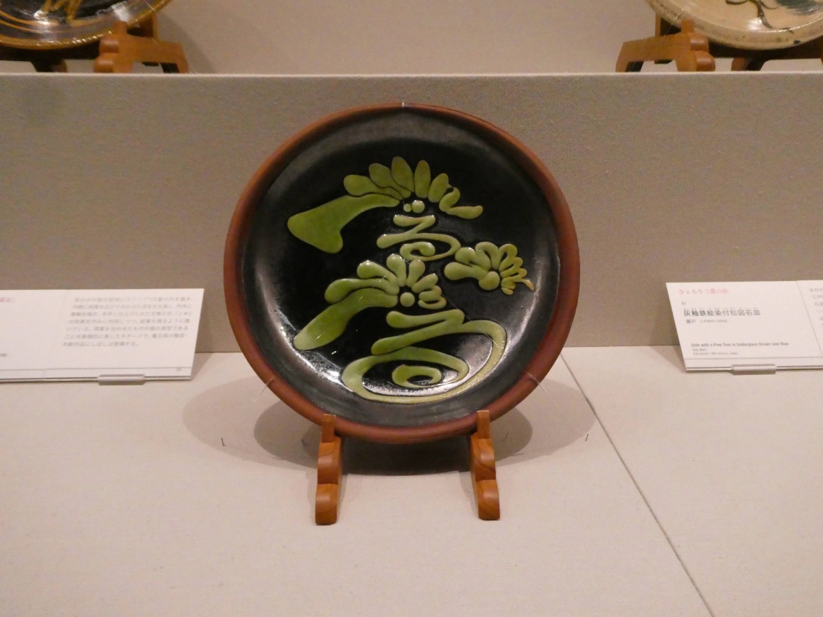 Japanese folk pottery dish with a green pine-tree design on black glaze, displayed on a wooden stand