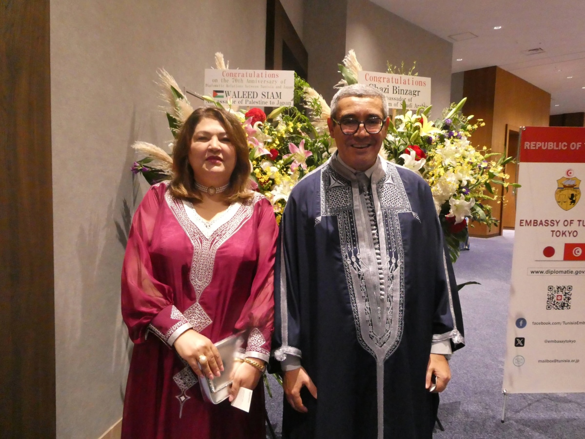Man and woman in traditional Tunisian dress in front of flower stands and the Embassy of Tunisia, Tokyo signboard