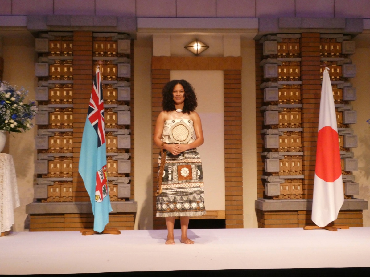 Woman in traditional Fijian attire standing between the Fijian and Japanese flags at the Fiji Day reception in Japan