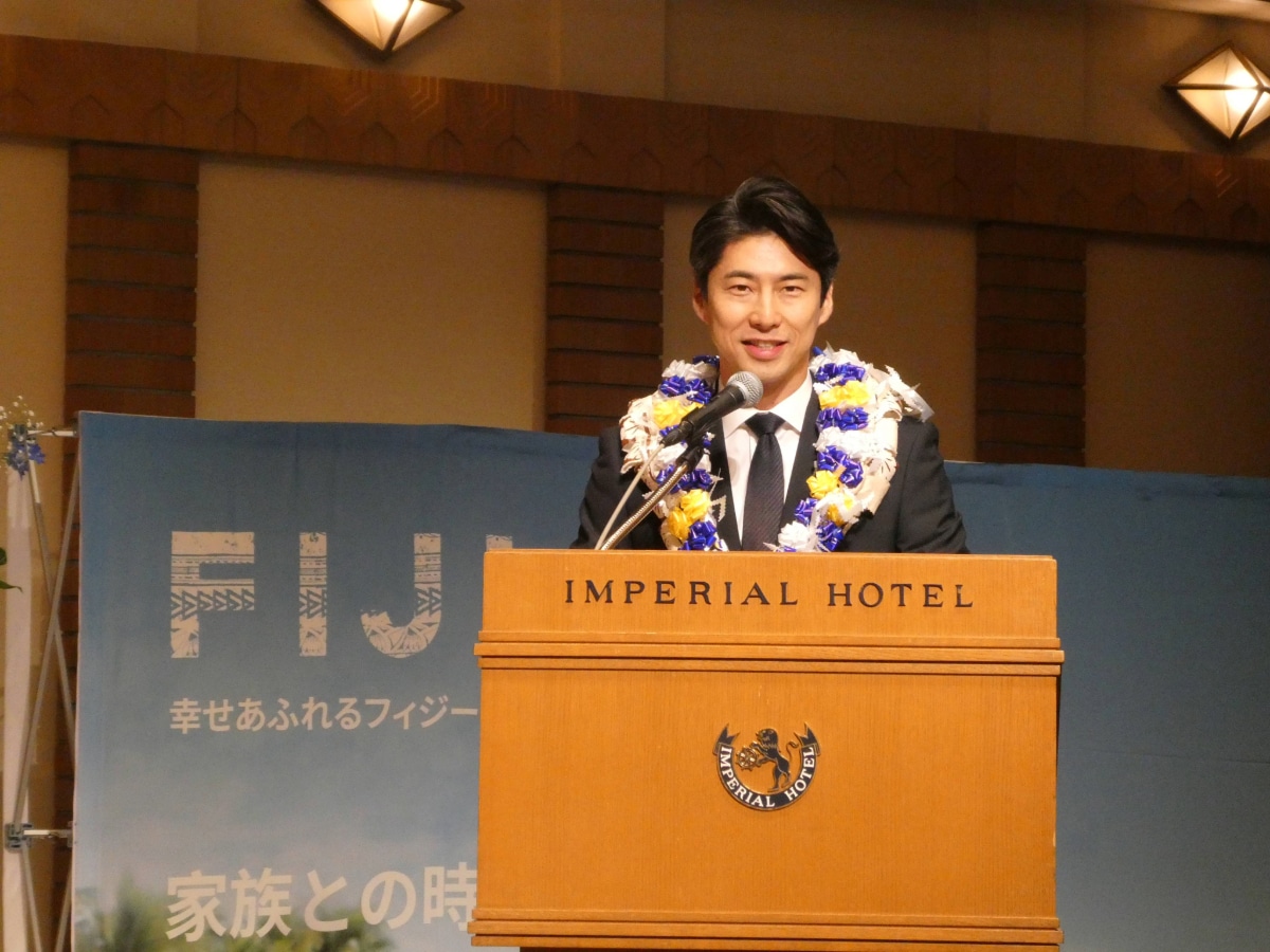 Man wearing a blue, yellow and white lei speaking at the podium during the Fiji Day reception in Japan