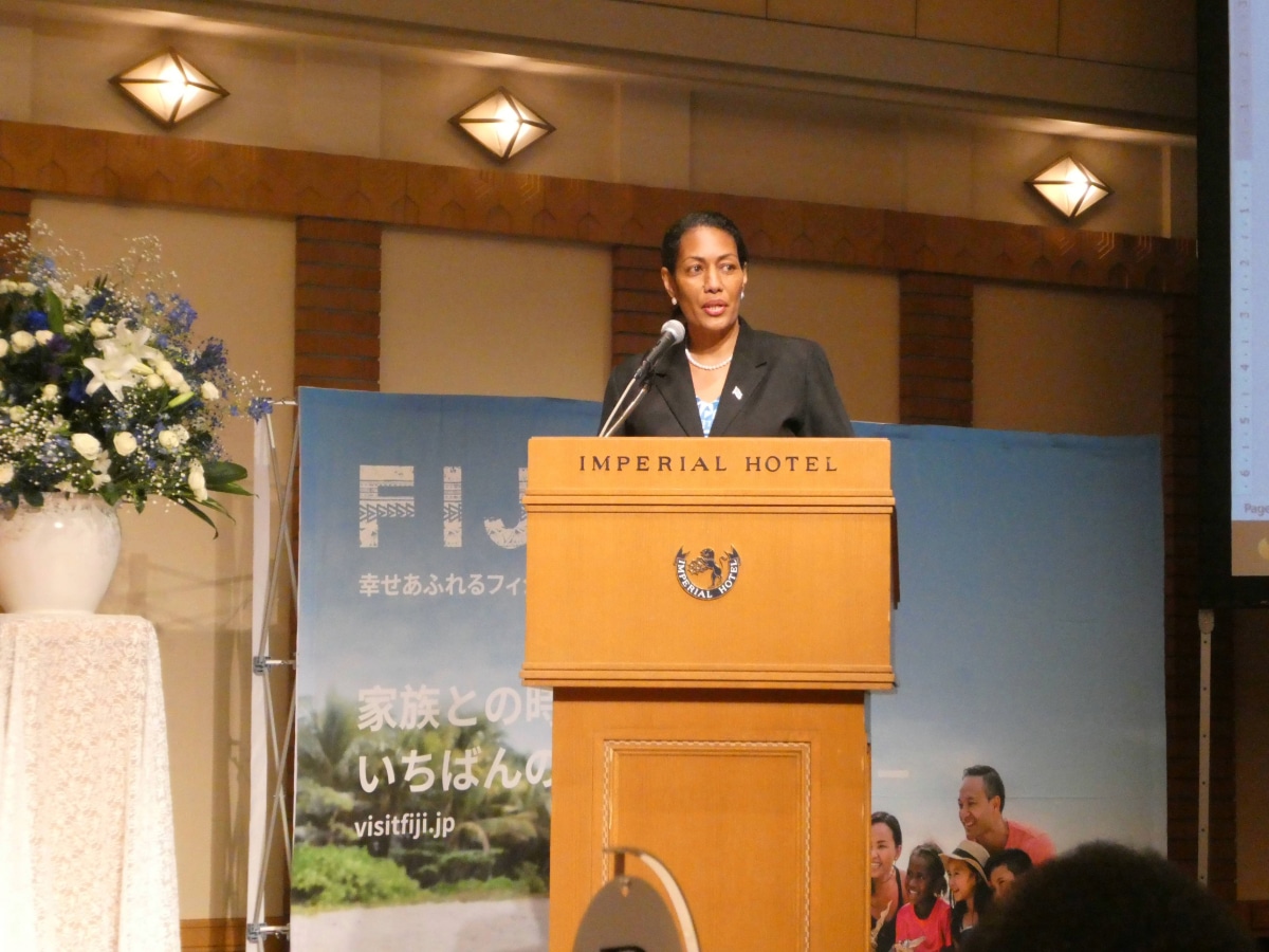 Woman delivering a speech at the podium during the Fiji Day reception, with a Fiji tourism banner behind her