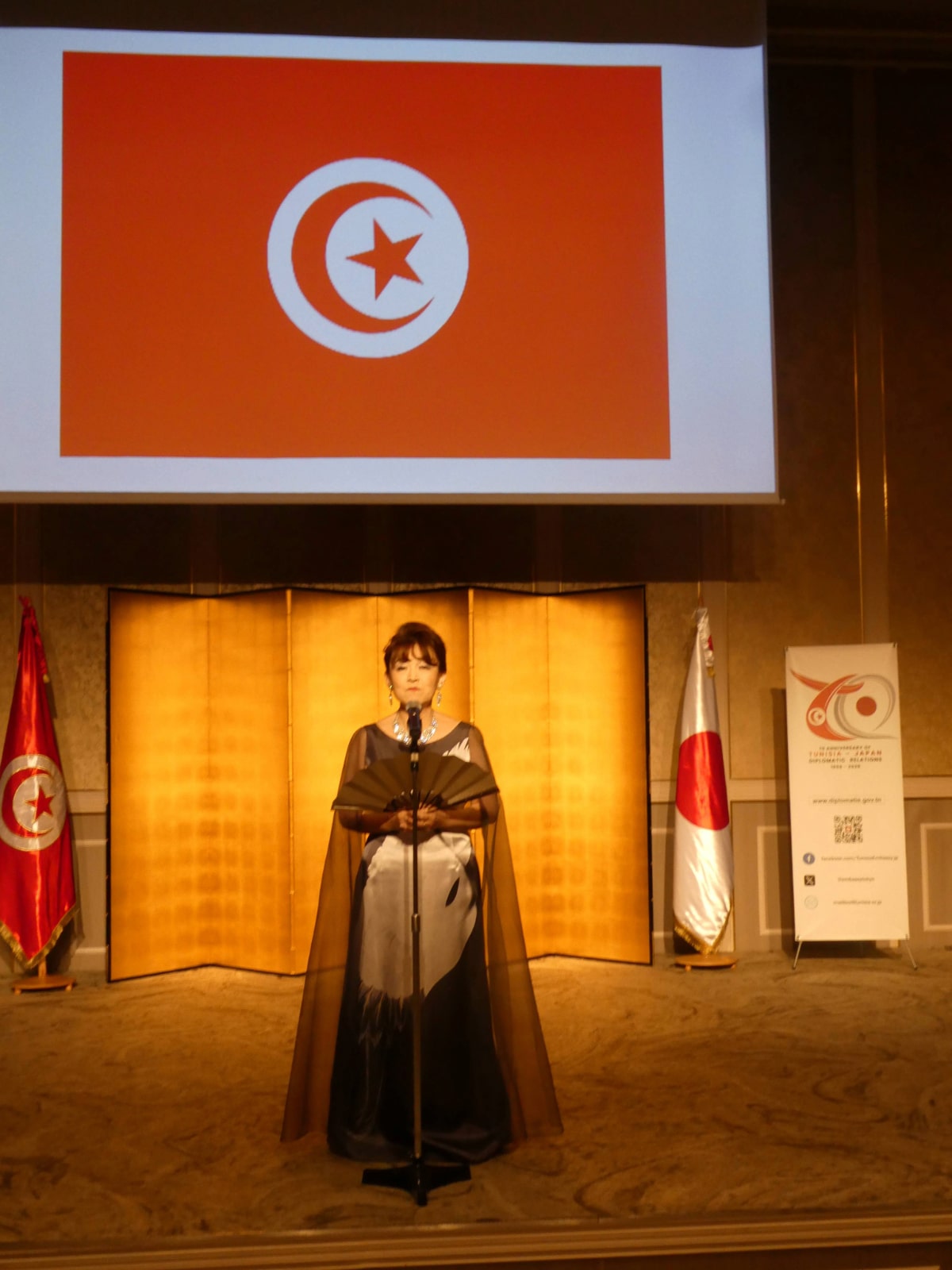 Woman in an evening gown holding a fan on stage with the Tunisian flag displayed on the screen above her