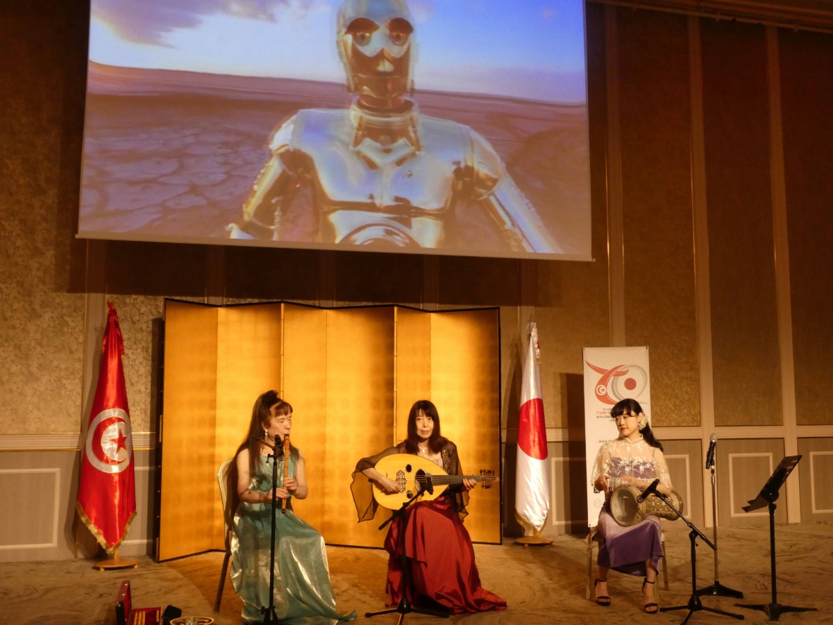 Three women playing flute, oud and drum on stage beside Tunisian and Japanese flags with a Star Wars scene on the screen