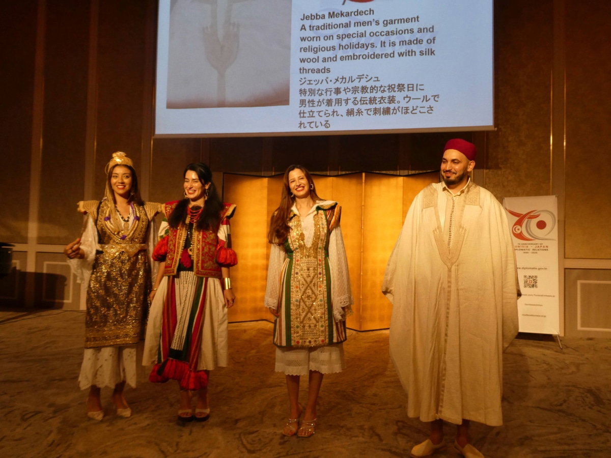 Models in traditional Tunisian costumes on stage in front of a slide describing the Jebba Mekardech men’s garment